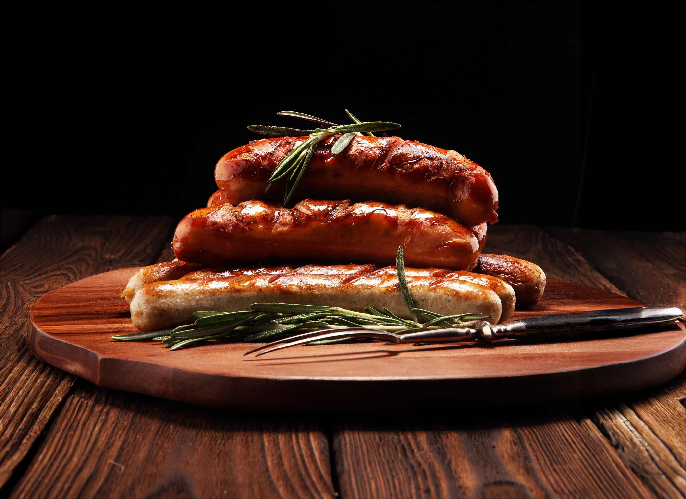 Gegrillte Würste auf einem Holzbrett, angerichtet mit Rosmarinzweigen und Grillbesteck, vor dunklem Hintergrund.