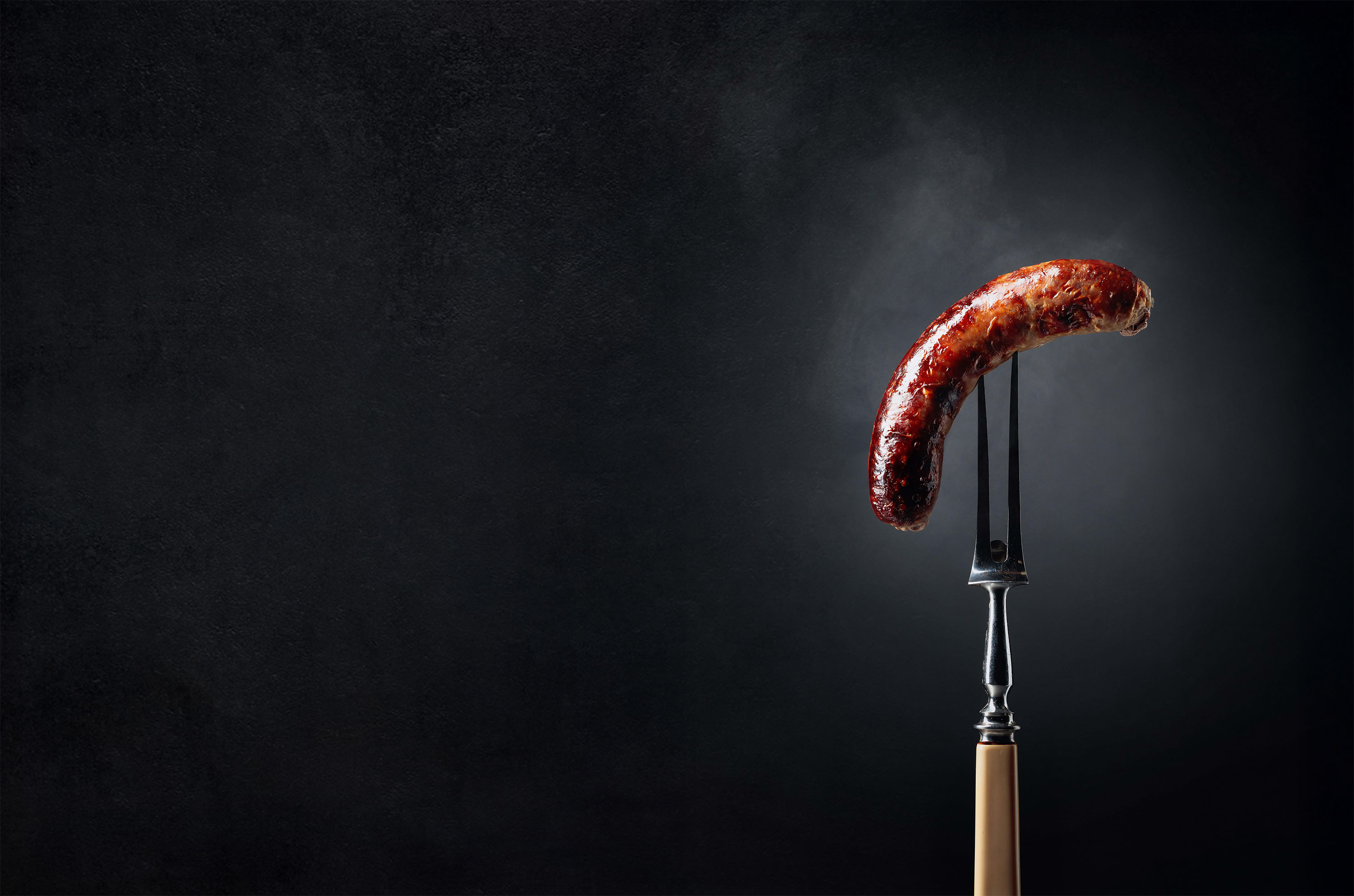 Gegrillte Wurst auf Fleischgabel vor dunklem Hintergrund